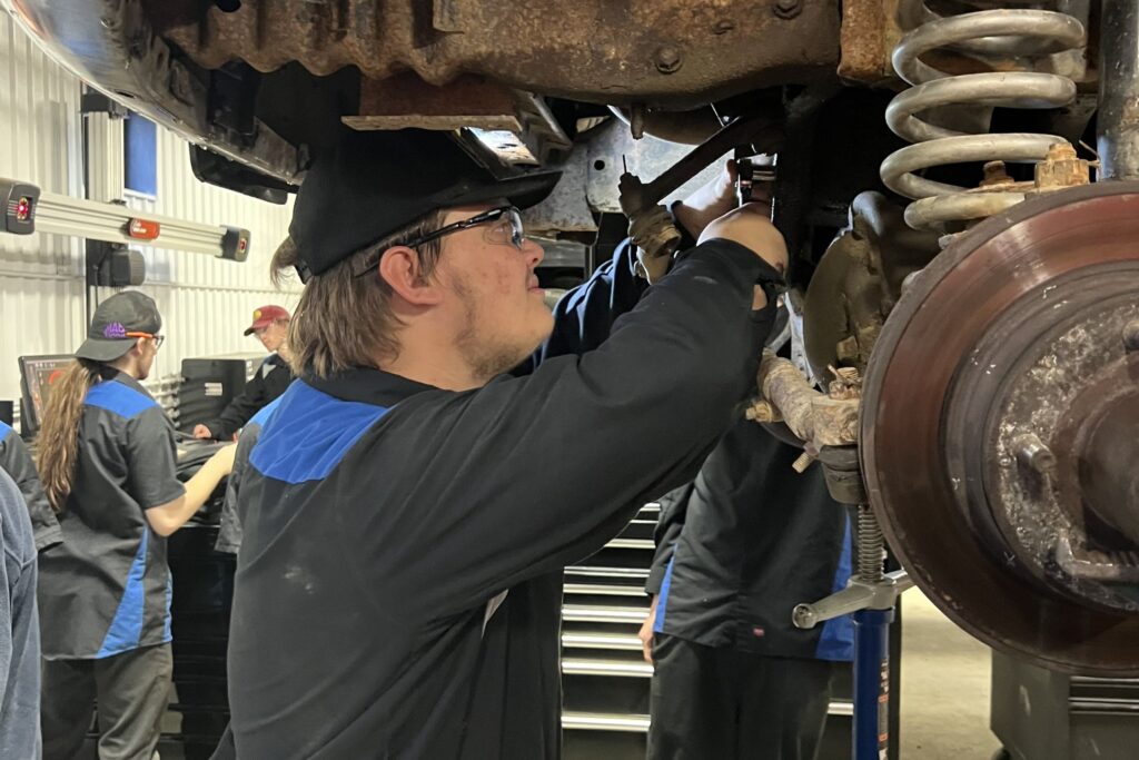 A male student in a ball cap stands to work underneath a car positioned on a lift in a class for an automotive career training program at a Western New York Works college