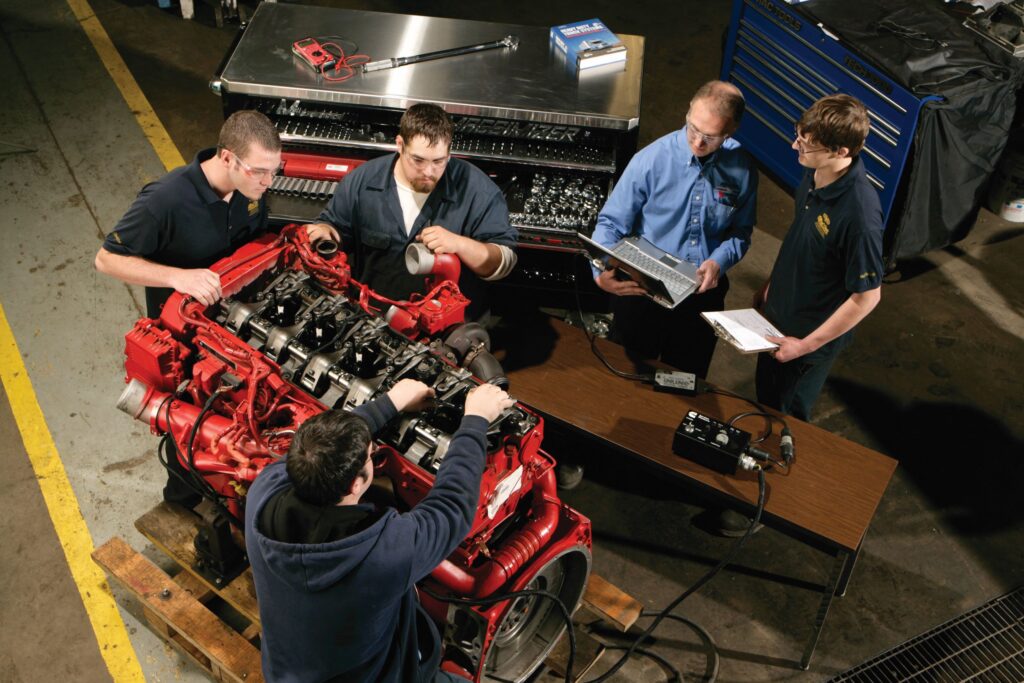 Four male students and an instructor holding a laptop explore the components of an engine in a class for a heavy equipment/diesel mechanic career training program at a Western New York Works college