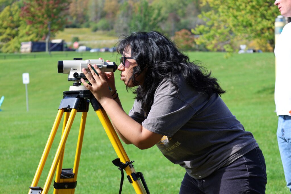 A female student wearing sunglasses practices using surveying equipment outdoors in a class for a construction career training program at a Western New York Works college.