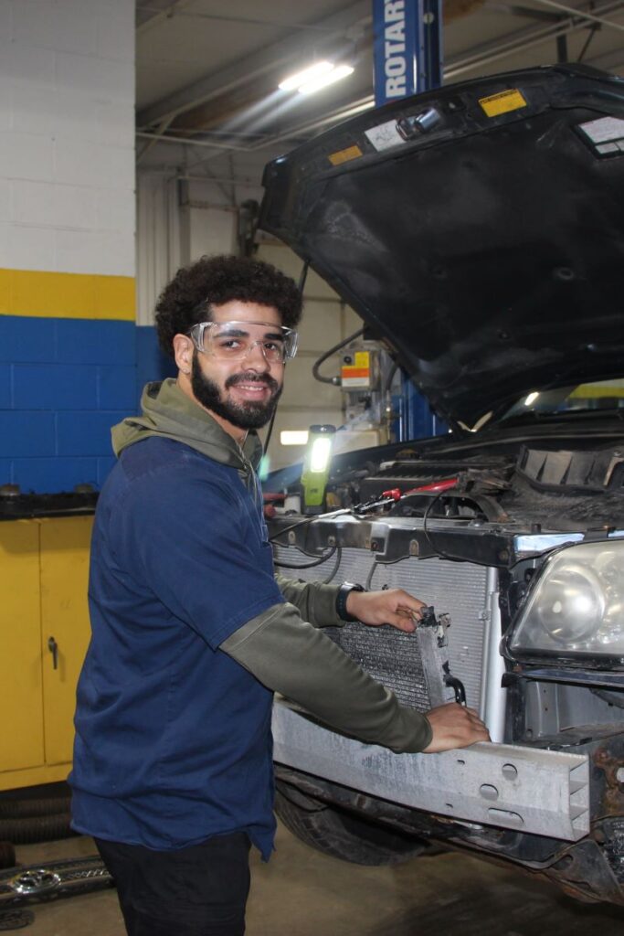 A male student wearing safety glasses stands in front of a vehicle with its hood open in a practice environment for Western New York Works' automotive career training programs