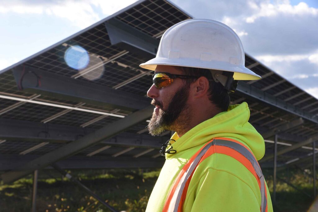 A student wearing a hard hat, reflective safety glasses, and a safety vest stands outdoors near solar panels in a class at a Western New York Works college.