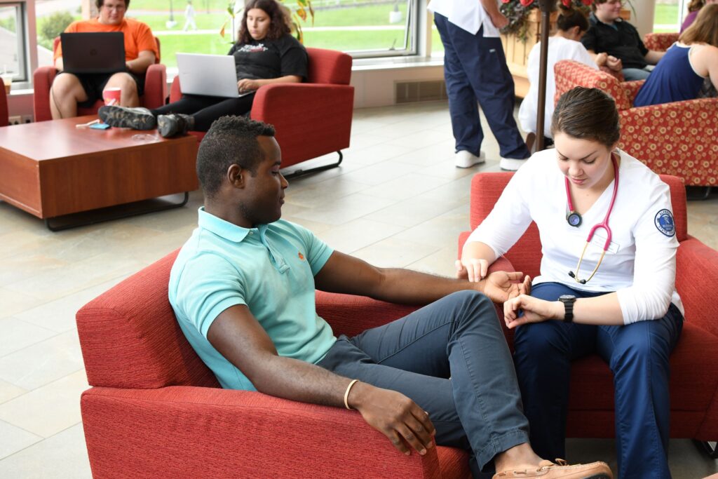 A student wearing a stethoscope looks at a watch while taking another student's pulse in a health information technician class at a Western New York Works college.