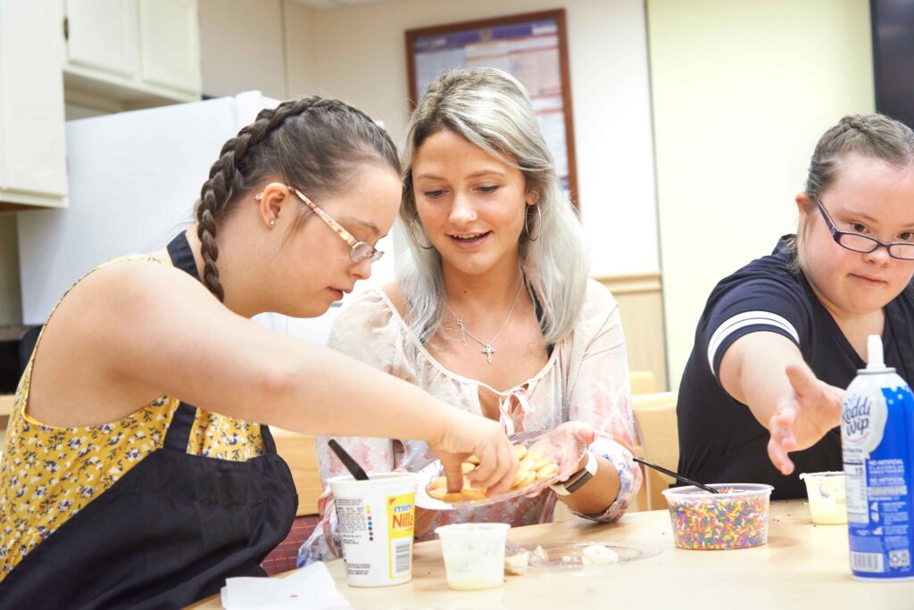 An occupational therapy assistant student at a Western New York Works college sits at a table with two patients who are reaching for potato chips and whipped cream.