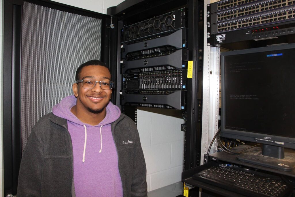 A male student stands by a keyboard and networking equipment in a computer repair technician career training program at a Western New York Works college.