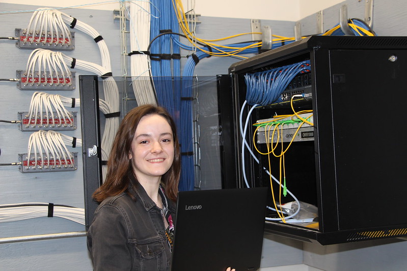 A student holding a laptop stands in front of a cabinet holding cables and networking equipment in a health information technician class at a Western New York Works college.