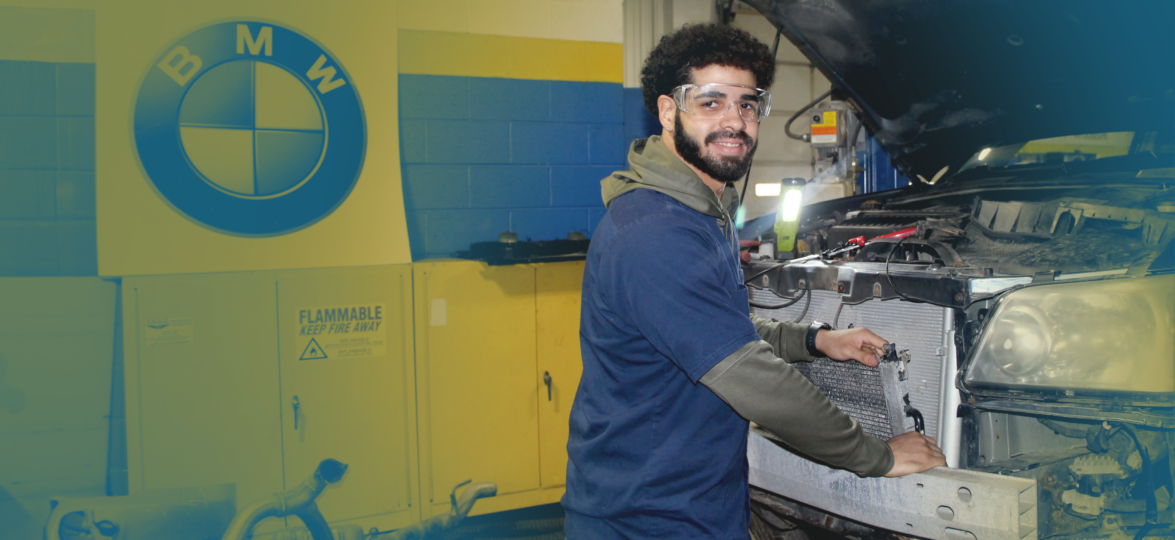 A male student wearing safety glasses stands in front of a vehicle with its hood open in a practice environment for Western New York Works' automotive career training programs